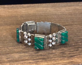 May include: Silver-toned bracelet with rectangular green accents and spherical silver details. The bracelet is made of multiple connected segments and is resting on a wooden surface.