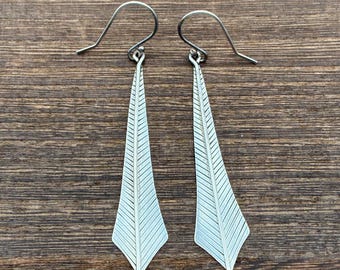 May include: A pair of silver-colored dangle earrings. Each earring features a long, triangular shape with etched linear details, suspended from a simple hook. The earrings are displayed against a wooden background.
