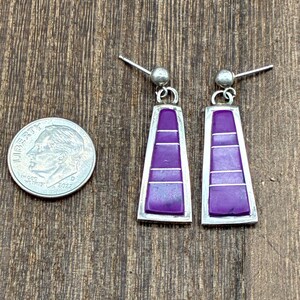 May include: A pair of silver-toned earrings with a rectangular, trapezoid shape. Each earring features a vibrant purple inlay with horizontal silver lines. The earrings are displayed next to a U.S. dime coin.