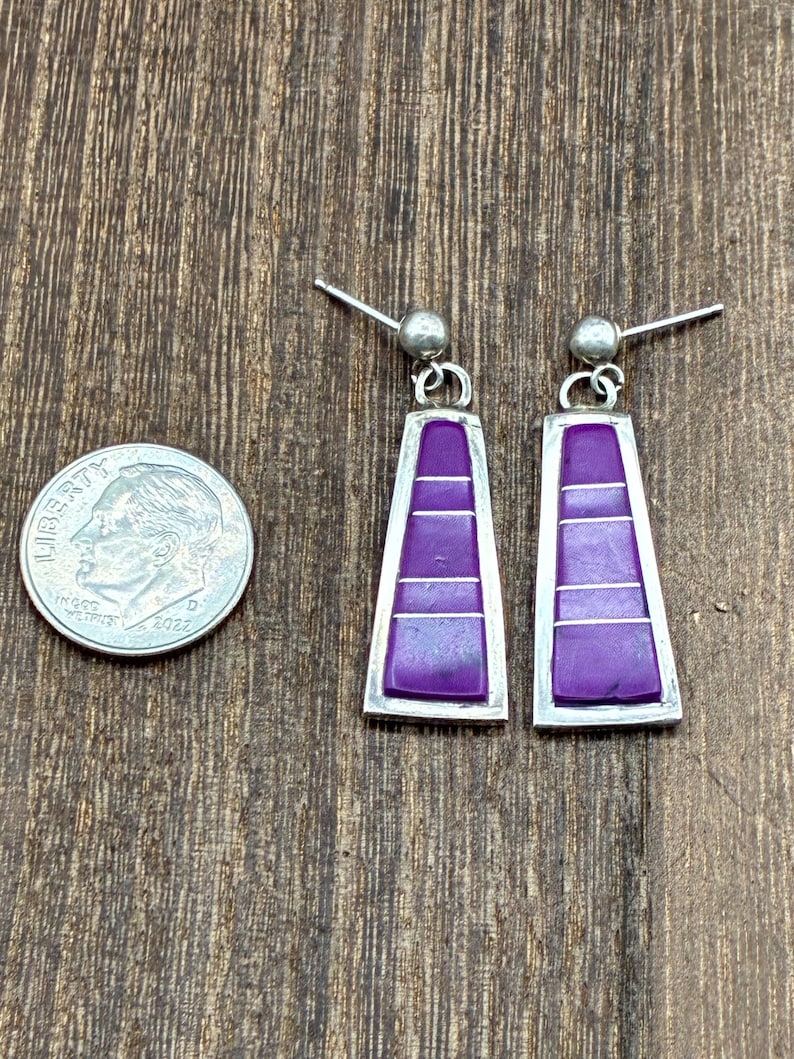May include: A pair of silver-toned earrings with a rectangular, trapezoid shape. Each earring features a vibrant purple inlay with horizontal silver lines. The earrings are displayed next to a U.S. dime coin.