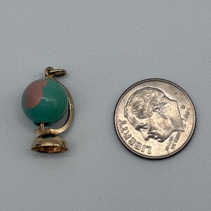 May include: A gold-toned globe charm featuring a teal and pink enameled sphere, positioned next to a United States dime for scale. The charm includes a small circular base and a curved support arm, with a jump ring attached at the top for hanging.
