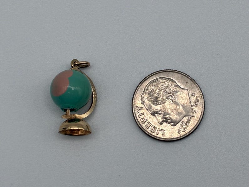 May include: A gold-toned globe charm featuring a teal and pink enameled sphere, positioned next to a United States dime for scale. The charm includes a small circular base and a curved support arm, with a jump ring attached at the top for hanging.