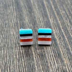 May include: Square stud earrings with a turquoise, black, red, and white striped design. The earrings are made of inlaid materials and have a silver post. The earrings are displayed on a wooden surface.