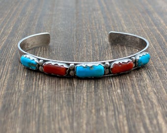 May include: A silver cuff bracelet featuring five rectangular, bezel-set stones arranged in an alternating pattern. The design includes three bright blue turquoise stones and two vibrant red coral stones, separated by small silver spheres.
