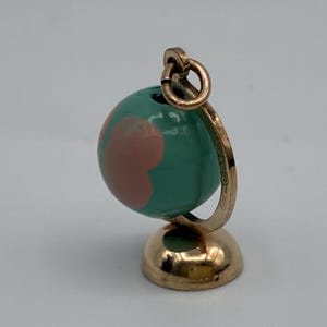 May include: A gold-toned metal charm shaped like a miniature globe. The sphere features a glossy teal surface with smooth, rounded pink shapes. It is mounted on a circular gold-toned base with a curved support arm and a small jump ring at the top.