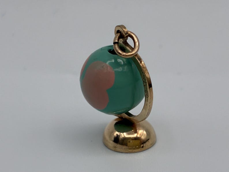 May include: A gold-toned metal charm shaped like a miniature globe. The sphere features a glossy teal surface with smooth, rounded pink shapes. It is mounted on a circular gold-toned base with a curved support arm and a small jump ring at the top.
