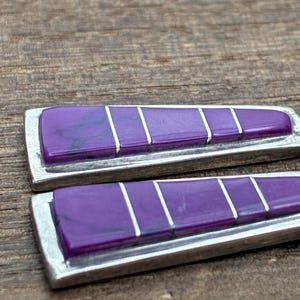 May include: Close-up of a pair of earrings with a rectangular design. Each earring features a vibrant purple stone with silver accents and a silver frame. The earrings are set against a wooden background.