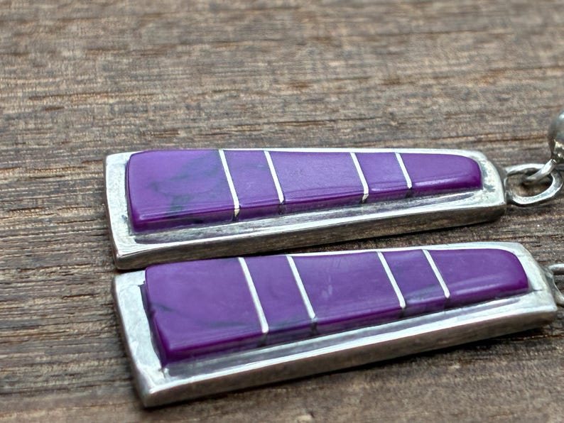 May include: Close-up of a pair of earrings with a rectangular design. Each earring features a vibrant purple stone with silver accents and a silver frame. The earrings are set against a wooden background.