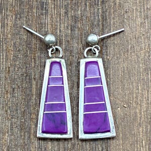 May include: A pair of silver-toned earrings with a geometric design. Each earring features a rectangular purple stone with three horizontal silver lines, set within a silver frame. The earrings have a post and ball closure.