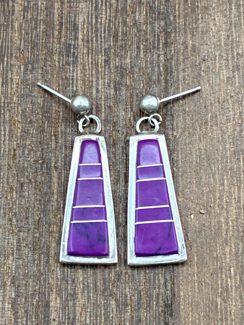 May include: A pair of silver-toned earrings with a geometric design. Each earring features a rectangular purple stone with three horizontal silver lines, set within a silver frame. The earrings have a post and ball closure.