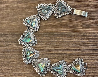 May include: Silver-toned bracelet featuring triangular abalone shell inlays. Each triangle is framed with a decorative silver border and linked together. The bracelet has a clasp closure and rests on a wooden surface.