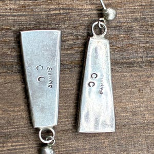 May include: A pair of silver earrings with a rectangular, tapered design. Each earring has a small ball at the bottom and a post for piercing. The word "Sterling" and the initials "CC" are visible on the back of each earring.