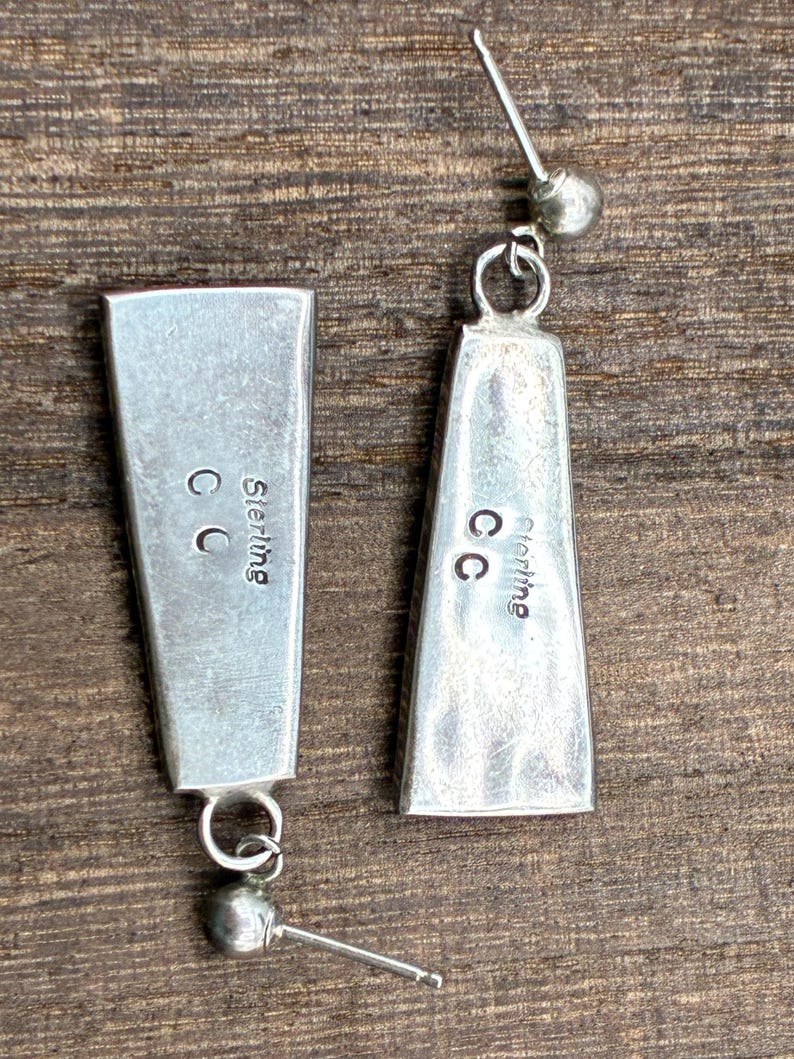 May include: A pair of silver earrings with a rectangular, tapered design. Each earring has a small ball at the bottom and a post for piercing. The word "Sterling" and the initials "CC" are visible on the back of each earring.