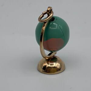 May include: A miniature globe charm featuring a metallic gold-toned stand and a circular hanging loop. The spherical globe is painted in shades of teal and muted pink, with a visible seam running vertically across the surface of the sphere.