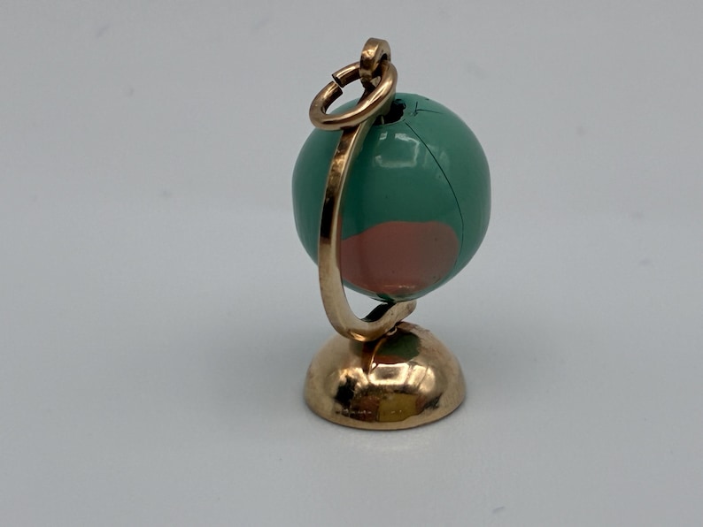 May include: A miniature globe charm featuring a metallic gold-toned stand and a circular hanging loop. The spherical globe is painted in shades of teal and muted pink, with a visible seam running vertically across the surface of the sphere.