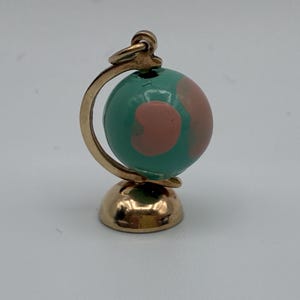 May include: A gold-toned metal charm shaped like a miniature globe. The sphere features a teal background with smooth, rounded pink shapes representing landmasses. It is mounted on a curved gold-toned stand with a circular base and a top loop.