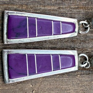 May include: A pair of silver-toned earrings with a geometric design. Each earring features a vibrant purple rectangular inlay divided by white lines, set within a silver frame. The earrings have a hook closure.