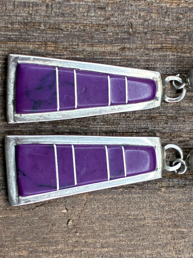 May include: A pair of silver-toned earrings with a geometric design. Each earring features a vibrant purple rectangular inlay divided by white lines, set within a silver frame. The earrings have a hook closure.