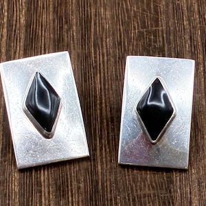 May include: A pair of rectangular silver cufflinks, each featuring a black diamond-shaped stone set in the center. The cufflinks are displayed on a dark wood surface, showcasing their polished metal and gemstone design.