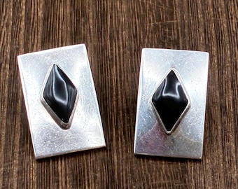 May include: A pair of rectangular silver cufflinks, each featuring a black diamond-shaped stone set in the center. The cufflinks are displayed on a dark wood surface, showcasing their polished metal and gemstone design.