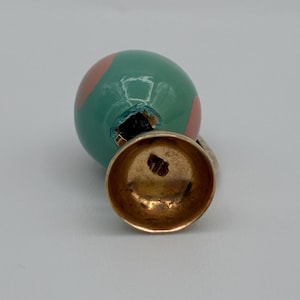May include: A spherical charm featuring a glossy teal surface with light pink circular accents. The charm is attached to a gold-toned metal bail with a hollow, bell-shaped base. The metal component shows a small, stamped hallmark on its interior.