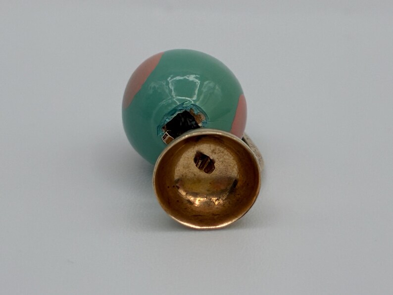 May include: A spherical charm featuring a glossy teal surface with light pink circular accents. The charm is attached to a gold-toned metal bail with a hollow, bell-shaped base. The metal component shows a small, stamped hallmark on its interior.