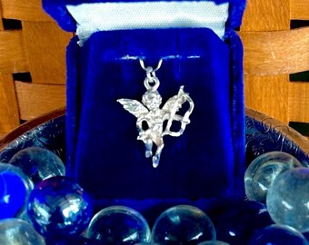 Puede incluir: Un amuleto de querubín de plata con alas y arco y flecha, presentado en una caja de terciopelo azul.