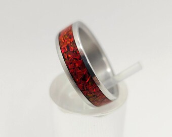 Puede incluir: Un anillo de plata con una incrustación roja y dorada. El anillo está en un expositor blanco.
