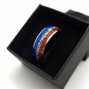 Può includere: Un anello d'argento con un'intarsio colorato di materiale blu, rosso e bianco simile all'opale. L'anello è presentato in una scatola nera, che mette in risalto il suo design vibrante. La fascia dell'anello è di colore argento lucido.