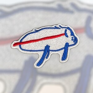 May include: Embroidered patch featuring a stylized white buffalo with blue legs and outline, and a red stripe across the body. The patch is likely for apparel or accessories, representing a sports team or organization.