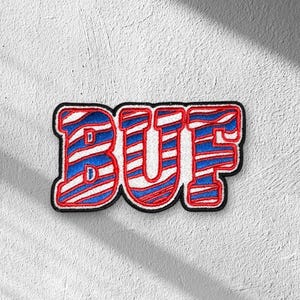 May include: An embroidered patch featuring the letters BUF in a bold, block font. The letters are filled with a repeating diagonal pattern of red, white, and blue stripes, and the entire design is outlined with a thick black border.