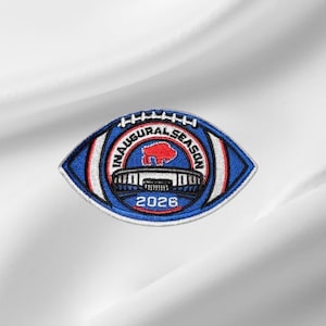 May include: An embroidered patch shaped like a football with blue, white, and red stitching. The center features a red buffalo silhouette above a stadium graphic, with the text INAUGURAL SEASON and the year 2026 printed in bold black lettering.
