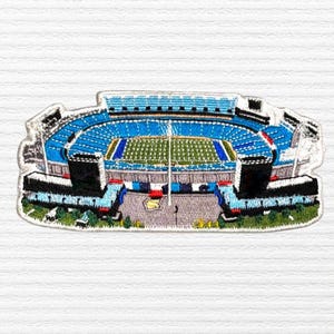 May include: Embroidered patch depicting a stadium with blue seating, a green field, and black structures. The patch has a white border and is set against a white background. The stadium features lights and a central goalpost.