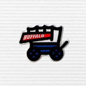 May include: Embroidered patch featuring a stylized covered wagon design. The wagon is black and white with blue accents and black wheels. The word "BUFFALO" is displayed in white letters on a red banner.