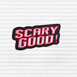 May include: A black and red embroidered patch with the words "SCARY GOOD!" in white, outlined in red. The patch has a black border and is set against a white, textured background. The design is bold and eye-catching.