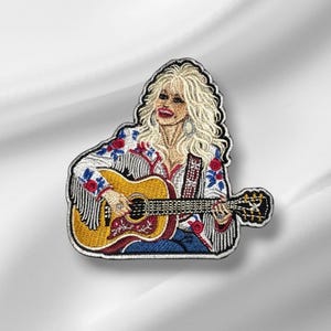 May include: An embroidered patch featuring a blonde person with long hair playing an acoustic guitar. The person wears a white jacket with red floral patterns and fringe details. The guitar is yellow with a dark fretboard and floral accents.