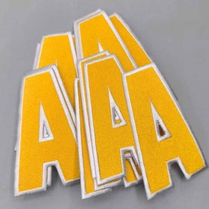 May include: A stack of yellow embroidered letter "A" patches with white borders. The patches are made of a textured fabric and are arranged on a gray surface. The letters are in a bold, block style.