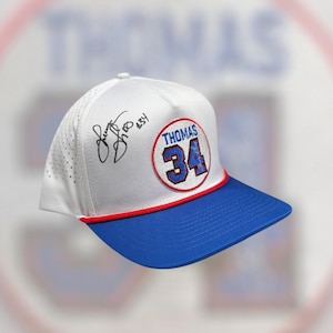 May include: White and blue baseball cap with a flat brim. The cap features a red and blue patch with the name "THOMAS" and the number "34". The cap has a blue brim and a white crown with a signature.
