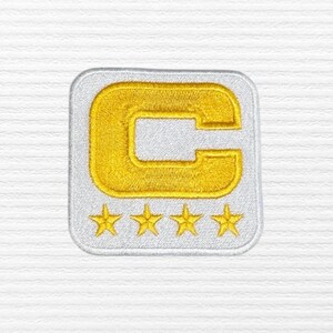 May include: Embroidered patch featuring a gold letter "C" inside a white square border. Below the letter are four gold stars. The patch is designed for application on clothing or accessories.