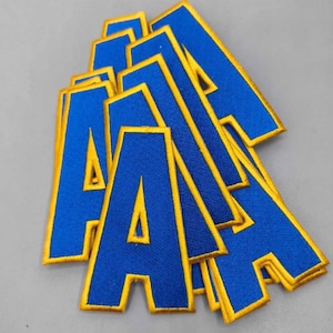 May include: A collection of blue fabric letter "A" patches with gold trim. The patches are stacked and slightly overlapping, creating a layered effect. The patches are likely for apparel or craft projects.