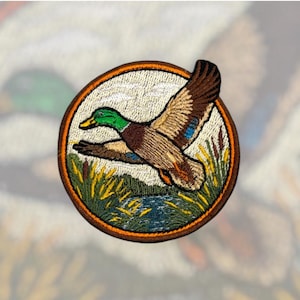 May include: Embroidered patch featuring a mallard duck in flight. The patch is circular, with a brown border and a cream-colored background. The duck has green, brown, and blue feathers, flying over a marsh scene with reeds and water.