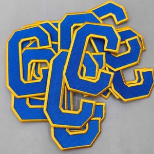 May include: A collection of blue fabric patches in the shape of the letter "C" with gold trim. The patches are scattered on a gray surface. The patches are likely for clothing or accessories.
