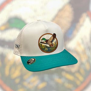 May include: A white and teal baseball cap with a detailed embroidered patch of a flying duck. The cap features the number "715" on the side and a teal brim. The duck patch is circular with a brown border.