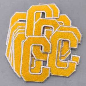 May include: A stack of yellow embroidered letter "C" patches with white borders. The patches are made of a textured fabric and are arranged on a gray surface. The patches are likely for apparel or crafts.