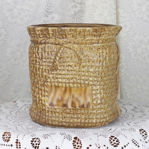 May include: A tan ceramic planter with a woven basket design. The planter has a rectangular panel on the front and a rope-like detail around the top. The planter is sitting on a white lace doily.