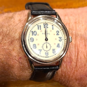 May include: A Fossil wristwatch with a cream-colored face, black numerals, and a small seconds dial. The watch has a silver-tone case and a black leather band. The time is approximately 12:00.