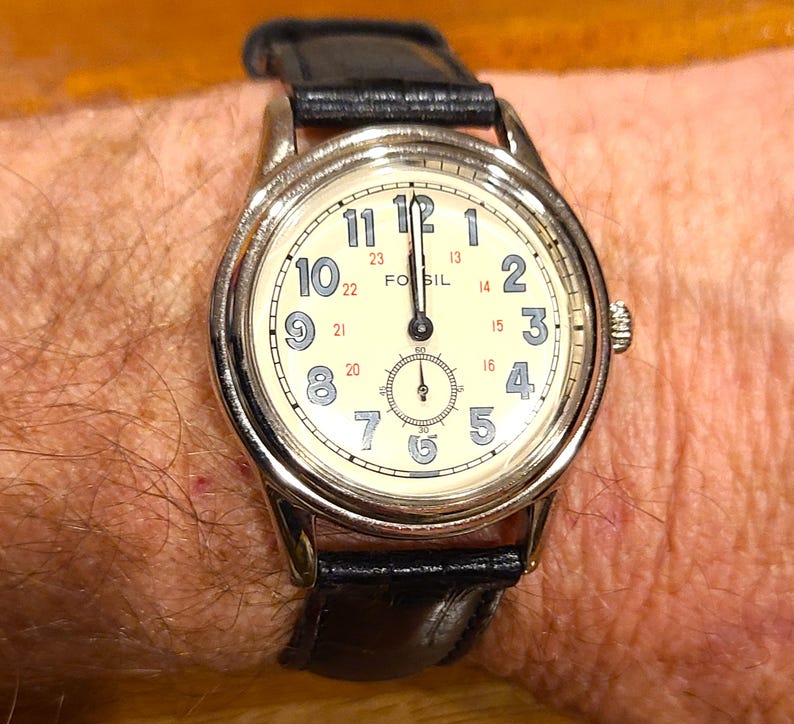 May include: A Fossil wristwatch with a cream-colored face, black numerals, and a small seconds dial. The watch has a silver-tone case and a black leather band. The time is approximately 12:00.