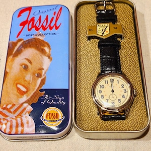 May include: A Fossil watch with a black leather band and a gold-toned case, displayed in a metal tin. The watch face is cream-colored with black numerals. The tin features the Fossil logo and a vintage image.