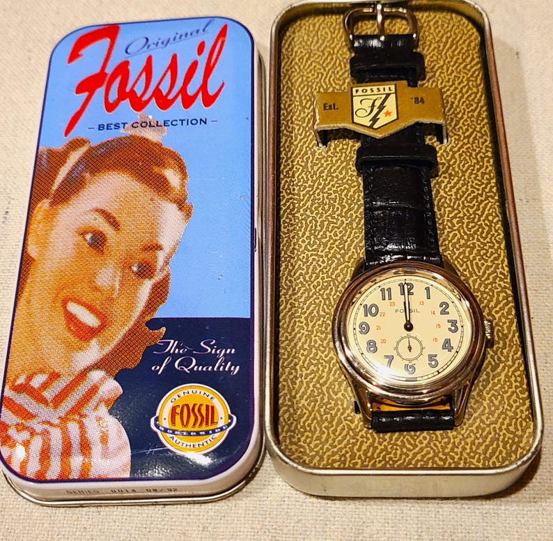 May include: A Fossil watch with a black leather band and a gold-toned case, displayed in a metal tin. The watch face is cream-colored with black numerals. The tin features the Fossil logo and a vintage image.
