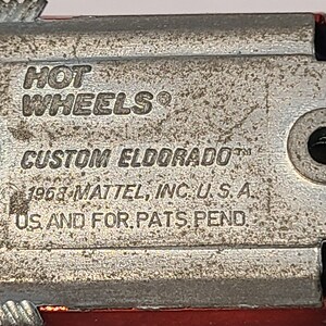 May include: Close-up of the underside of a red toy car with the text "HOT WHEELS" and "CUSTOM ELDORADO" printed on the metal chassis. The text "1968-MATTEL, INC. U.S.A." and "US AND FOR.PATS.PEND." are also visible.